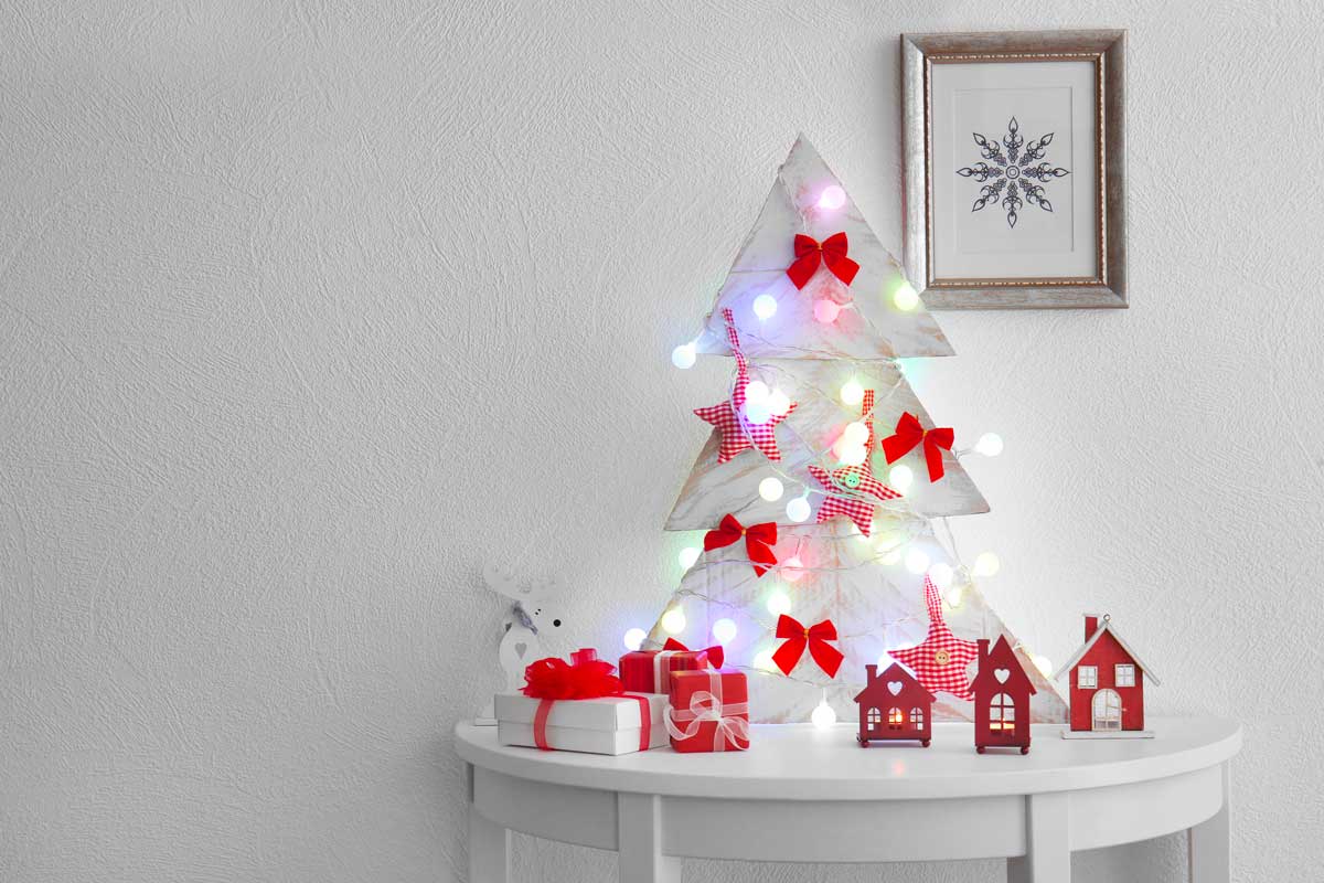 Alberello alternativo di Natale realizzato con pallet sbiancato, decorato con luci colorate, fiocchi rossi e stelle di stoffa, su tavolino bianco con pacchetti regalo e casette rosse.