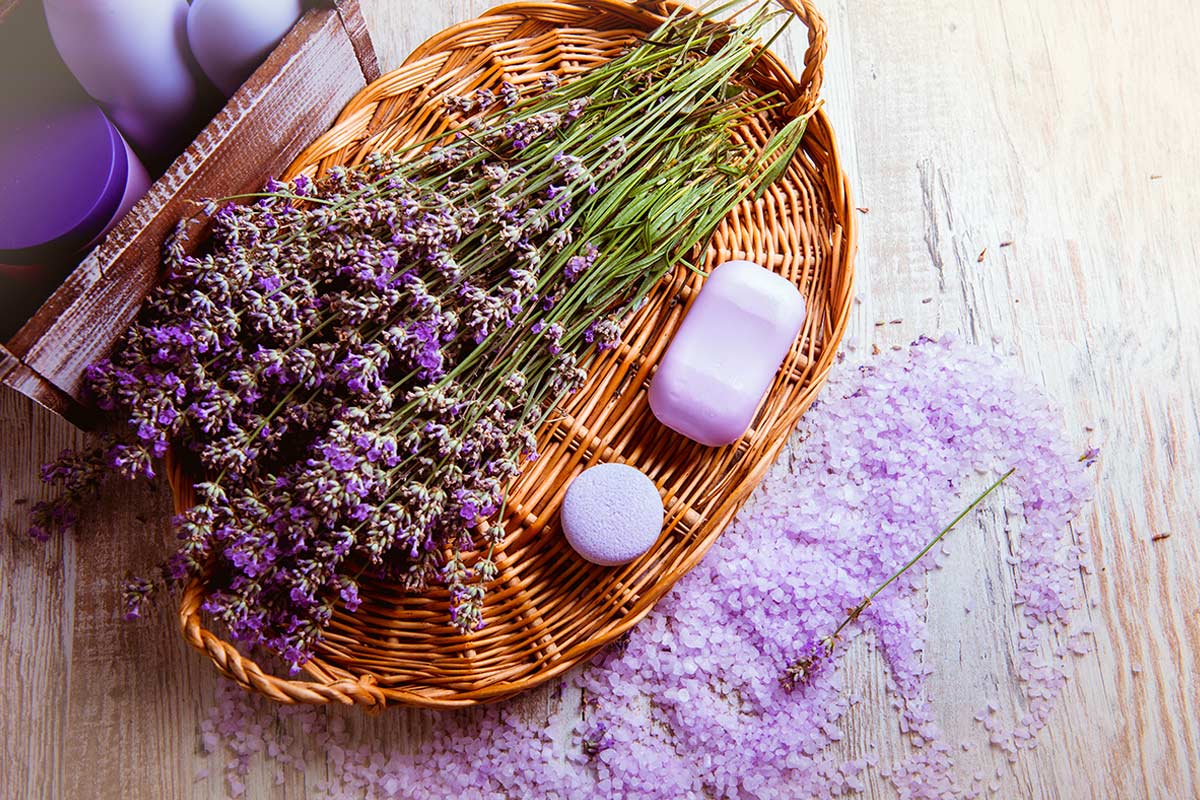 Cestino di vimini con mazzo di lavanda essiccata, sapone lilla e sali da bagno color lavanda sparsi su un tavolo in legno chiaro, atmosfera shabby e rilassante