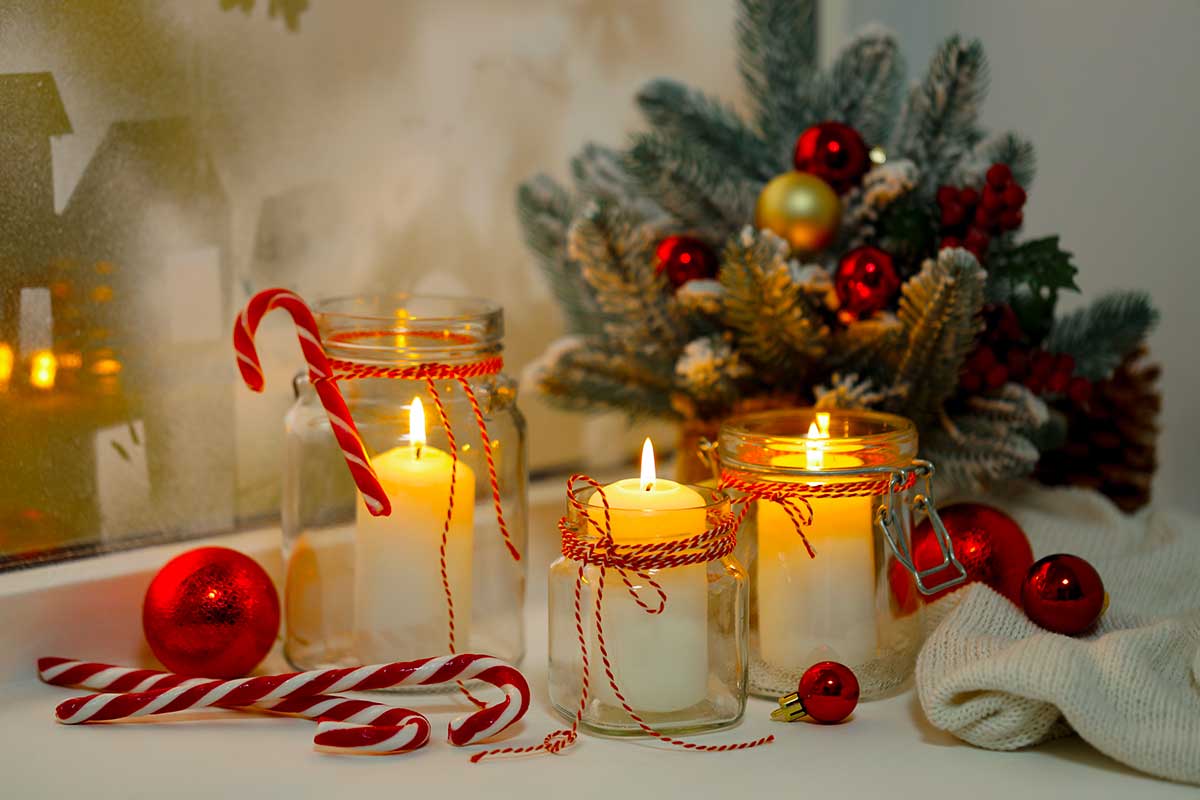 Barattoli di vetro con candele accese, bastoncini di zucchero e decorazioni natalizie rosse su una superficie chiara, con rametti di pino e palline invernali sullo sfondo.
