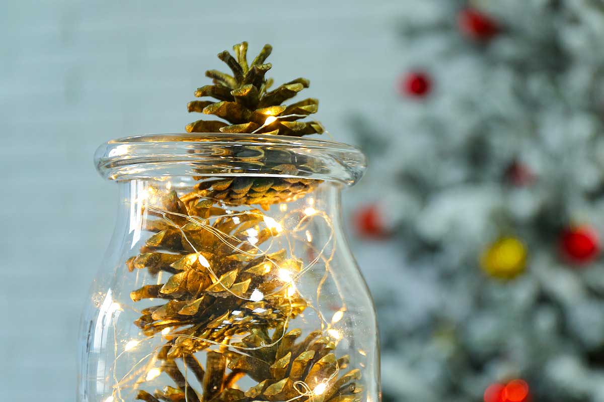 Barattolo di vetro con pigne dorate e lucine a led, in primo piano, su sfondo sfocato con albero di Natale decorato.