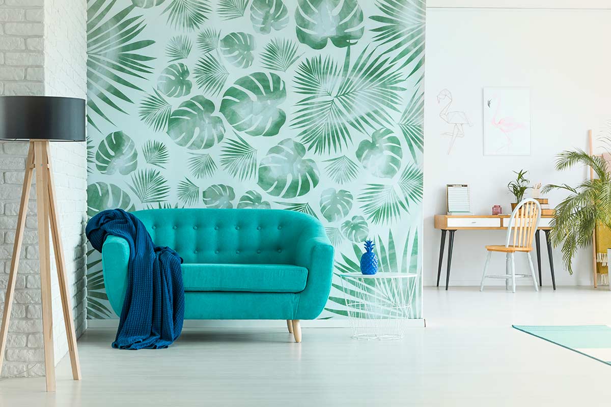 Parete decorata con carta da parati a motivo tropicale verde, divano azzurro e arredi moderni in un soggiorno luminoso e fresco. L’atmosfera naturale è accentuata da dettagli vegetali e colori vivaci, con angolo studio sullo sfondo.