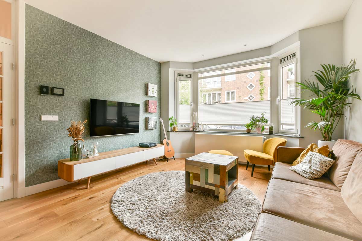 Soggiorno moderno con carta da parati verde a motivi delicati, pavimento in legno chiaro, ampio divano beige, grandi finestre luminose e piante d’arredo