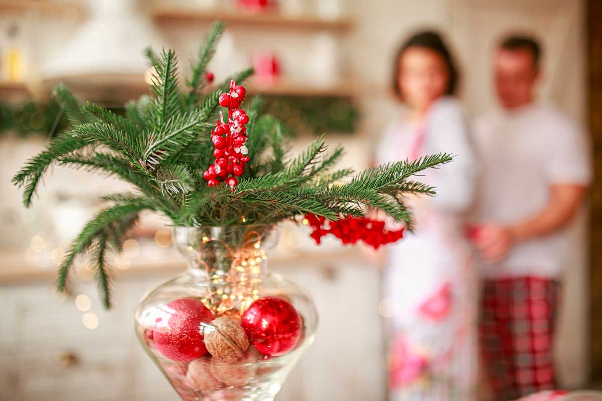 Vaso di vetro con rami di pino, bacche rosse, noci e palline natalizie rosse, decorato da lucine calde. Sullo sfondo, una coppia sfocata in cucina crea un’atmosfera accogliente e festiva.