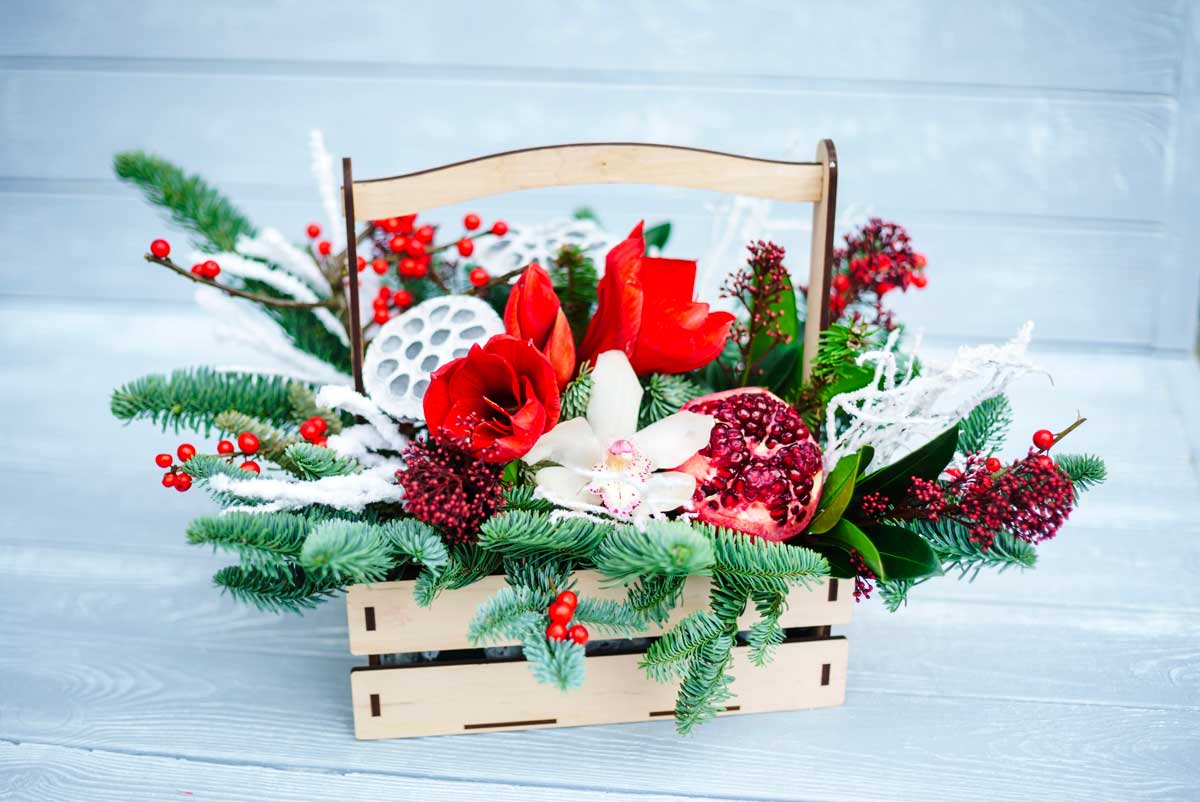 Cestino in legno con composizione natalizia di rami di abete, bacche rosse, fiori freschi, melagrana aperta e decorazioni bianche. Atmosfera vivace e naturale per le feste.