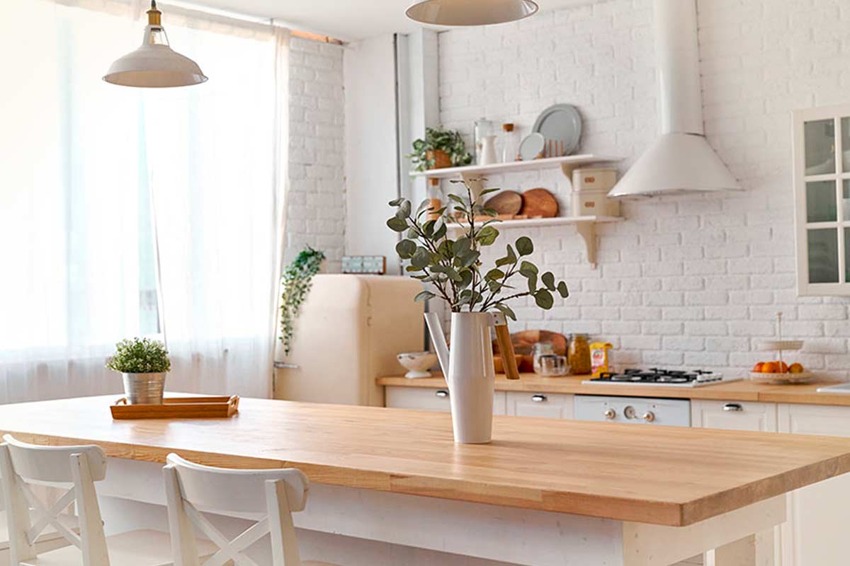 Cucina luminosa in stile nordico, con tavolo in legno chiaro, sedie bianche e dettagli naturali. Sul tavolo, un vaso con rami di eucalipto aggiunge un tocco verde fresco. Sullo sfondo, parete in mattoni bianchi, mensole con piatti e accessori, frigorifero retrò color panna e luce naturale che filtra da tende leggere. Atmosfera accogliente e ordinata.