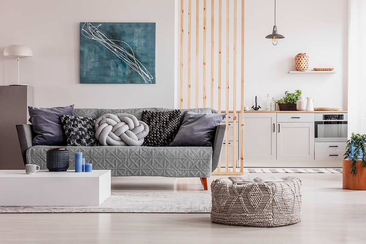 Soggiorno moderno e luminoso integrato con cucina bianca, divano grigio con cuscini decorativi, pouf lavorato a maglia, separé in legno chiaro e dettagli naturali che scaldano l’ambiente.