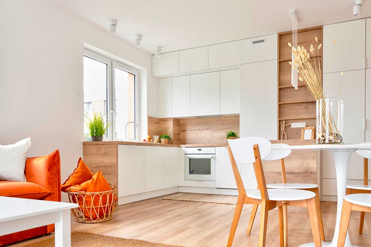 Cucina minimal bianca con dettagli in legno chiaro, arredi essenziali e luci calde. Tavolo e sedie in legno naturale, piante aromatiche e cuscini color arancio creano un’atmosfera luminosa e accogliente.