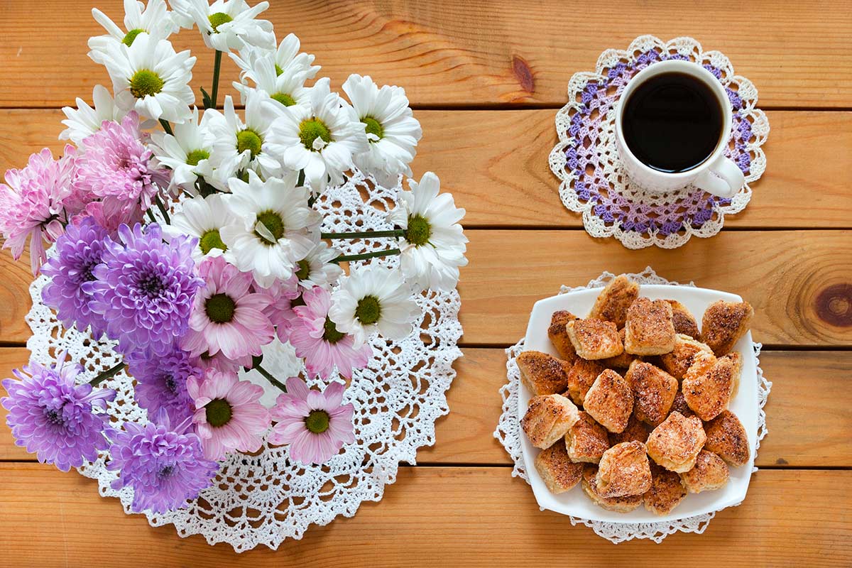 Tavolo in legno naturale con centrini all’uncinetto bianchi e lilla. Un mazzo di margherite bianche e crisantemi lilla in vaso, una tazza di caffè poggiata sul centrino, e un piatto di biscotti dorati, per una colazione dal sapore vintage e artigianale in stile shabby chic.