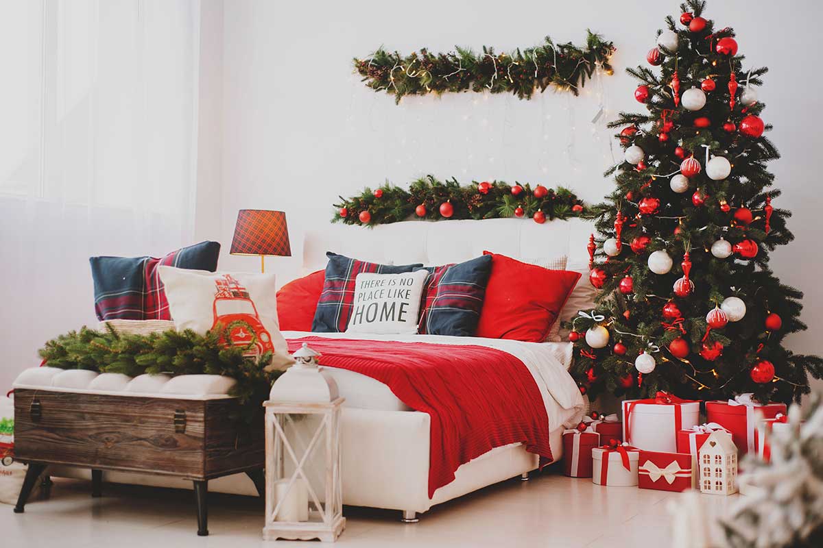 Camera da letto decorata per Natale con albero pieno di palline rosse e bianche, letto bianco con cuscini scozzesi e plaid rosso, festoni verdi e tanti pacchetti regalo. Stile caldo e accogliente.