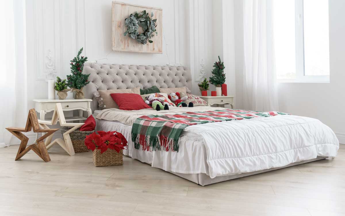 Camera da letto luminosa con letto bianco, plaid scozzese, cuscini rossi e decorazioni verdi. Stella di legno e poinsettia rossa completano l’atmosfera natalizia naturale.
