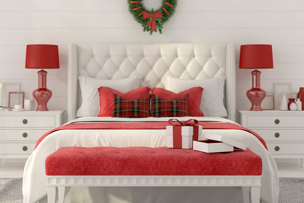 Camera da letto elegante con dettagli rossi, letto bianco, cuscini tartan, ghirlanda sopra la testiera e pacchetti regalo su una panca ai piedi del letto. Atmosfera raffinata e festosa.