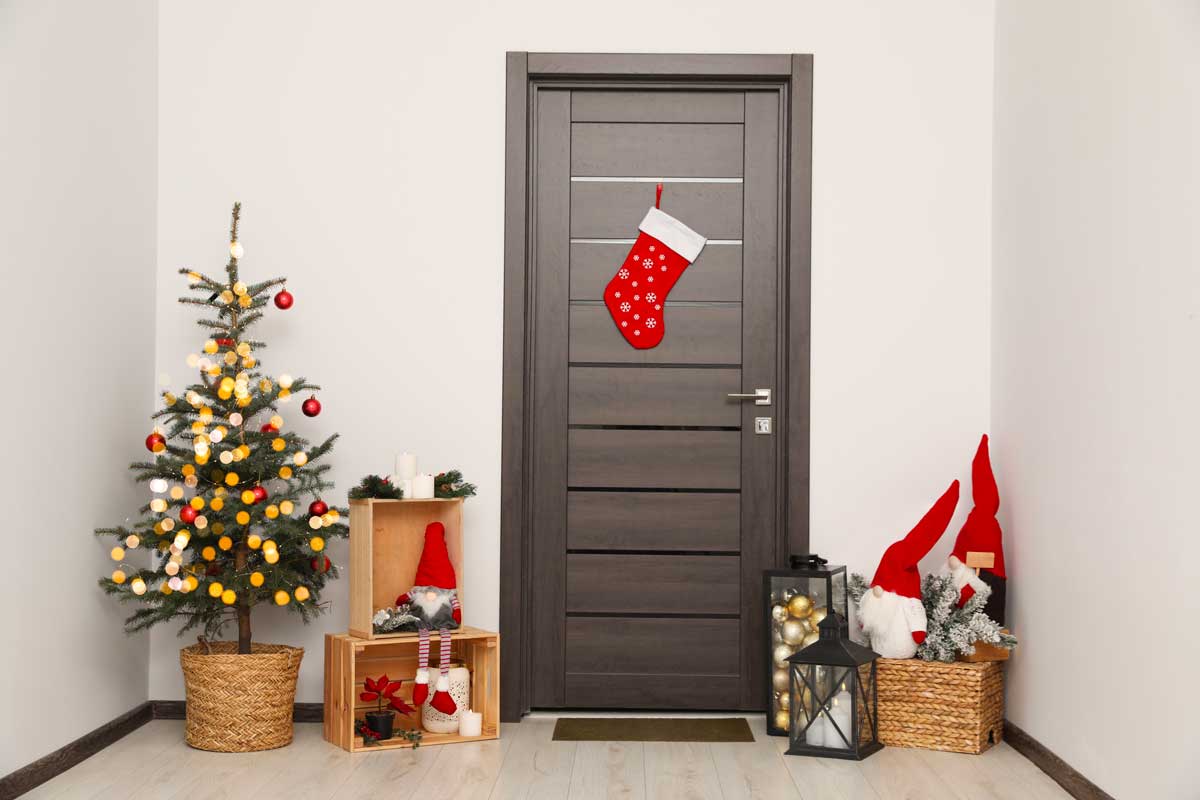 Ingresso interno decorato per Natale con albero, lanterna, calza rossa e addobbi festivi vicino a una porta moderna.