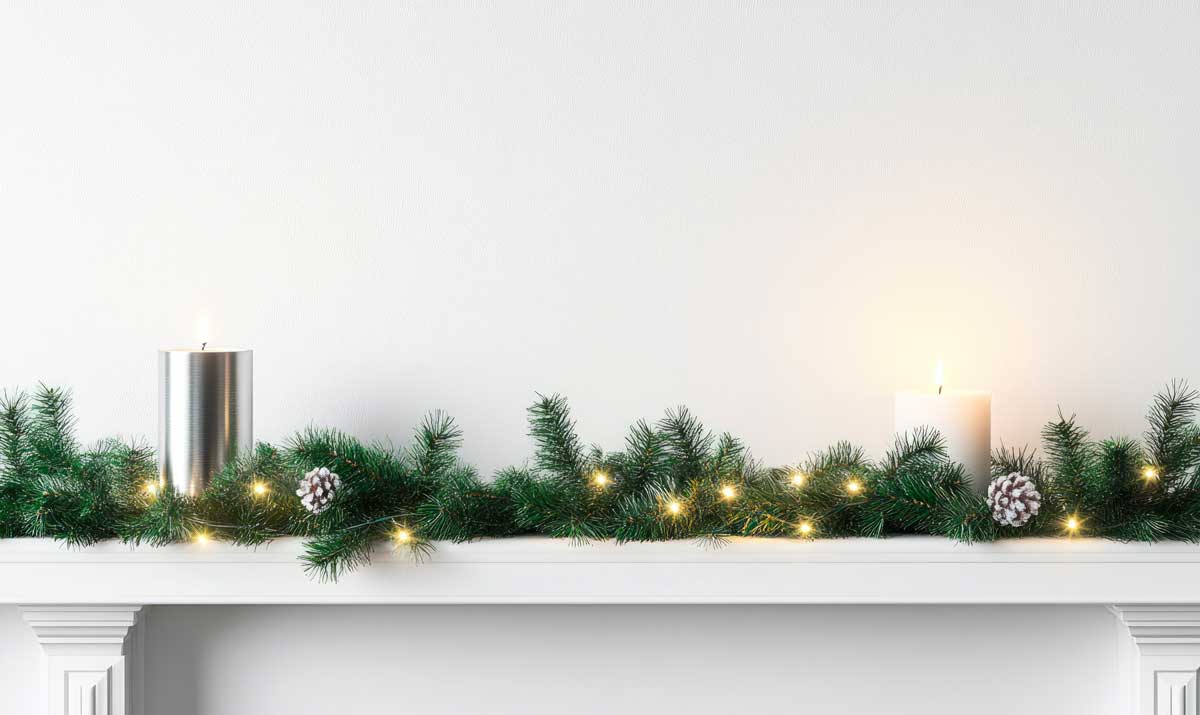 Mensola bianca decorata per Natale con una ghirlanda di rami di pino, pigne innevate, luci calde e due candele accese, su sfondo chiaro minimalista.