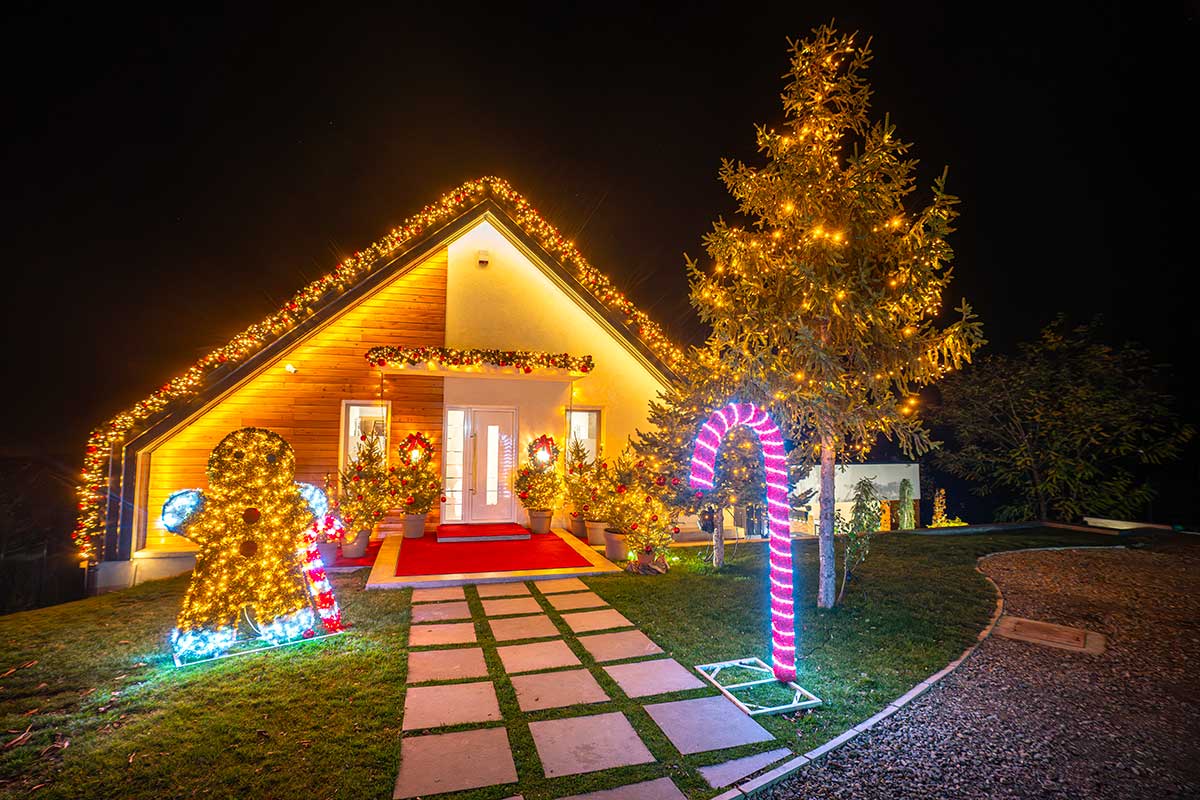 Casa moderna decorata per Natale con ghirlande luminose sul tetto, alberelli e corone illuminate, figure giganti di pan di zenzero e bastoncino di zucchero colorato sul prato. Atmosfera festosa e creativa nella notte.