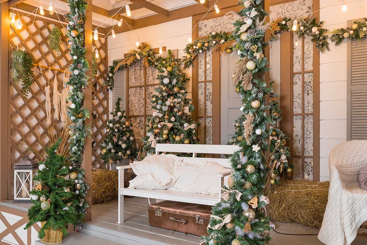 Veranda decorata per Natale con ghirlande di abete, luci calde e alberelli adornati con palline oro e bianche. Una panchina bianca con cuscini accoglie gli ospiti, mentre dettagli rustici e naturali creano un’atmosfera accogliente e festosa.