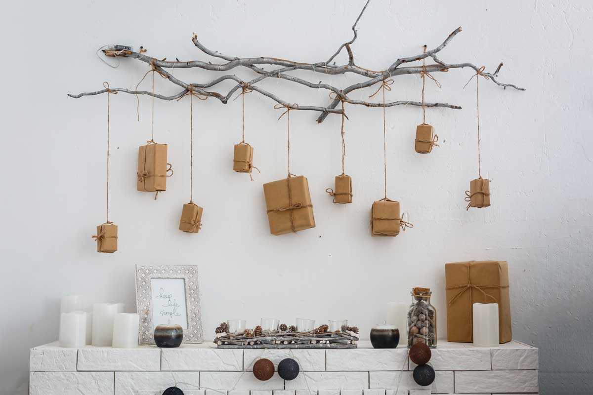 Ramo naturale appeso al muro con pacchetti regalo in carta kraft sospesi a diverse altezze con spago, sopra una mensola decorata con candele bianche, barattoli e oggetti naturali.