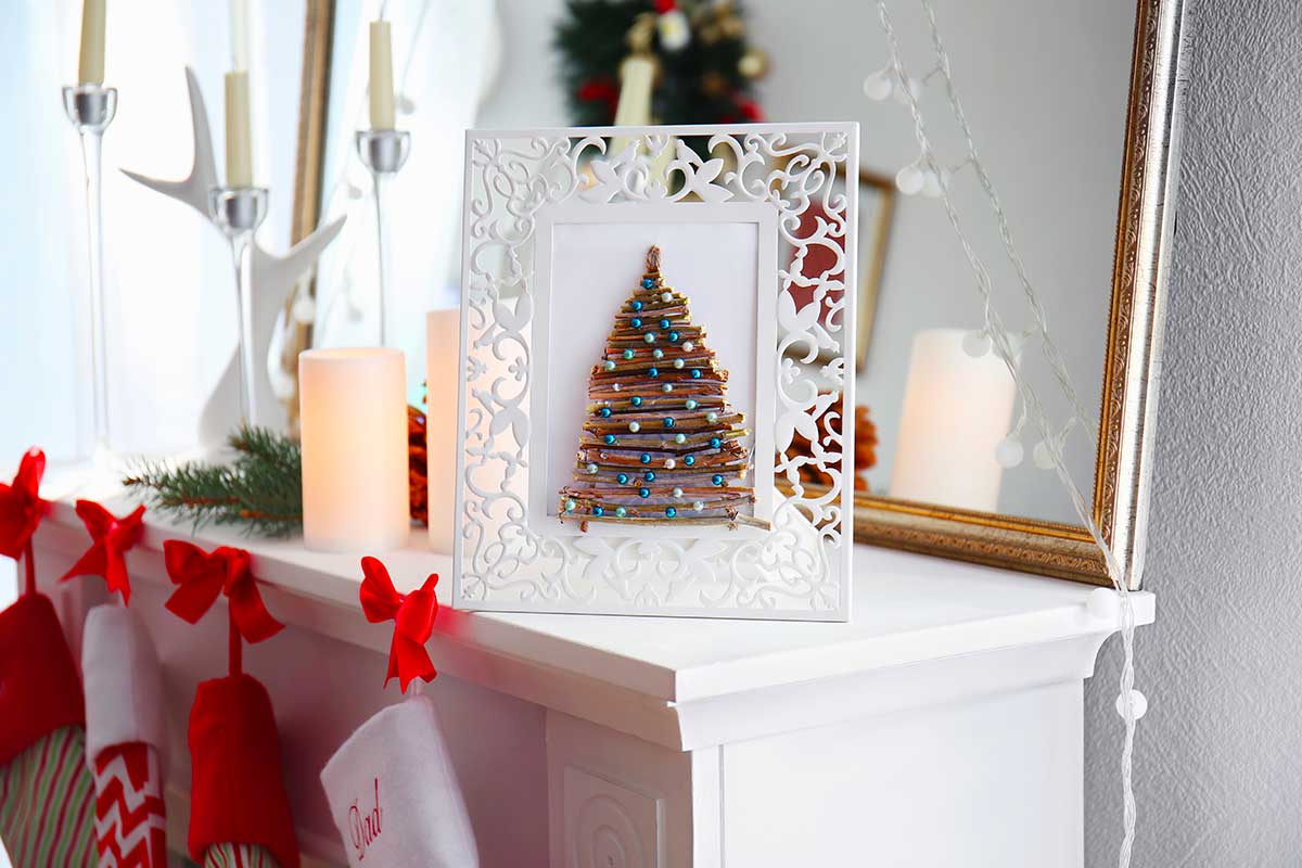 Cornice decorativa con al centro un albero di Natale stilizzato realizzato con piccoli rametti e perline colorate, appoggiata su un caminetto bianco decorato con candele, calze rosse e dettagli naturali.