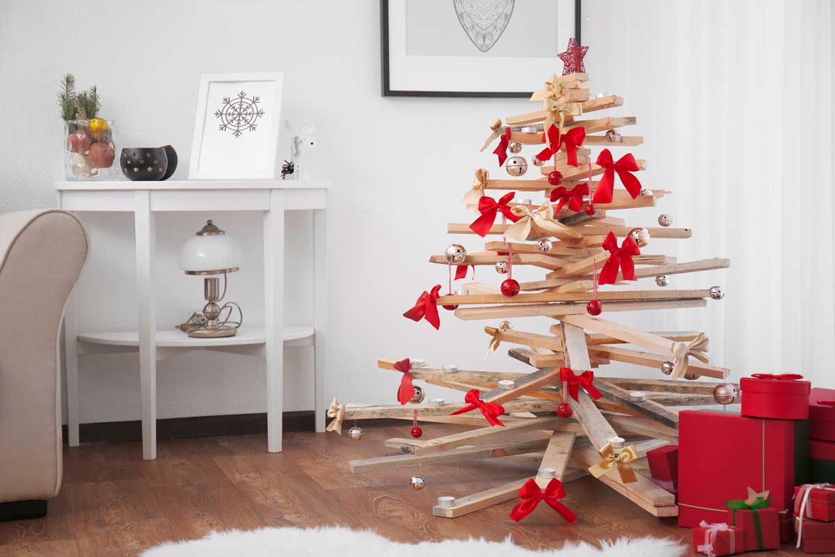 Albero di Natale realizzato con assi di legno naturale, decorato con fiocchi rossi, palline dorate e rosse, in un salotto moderno con pacchi regalo rossi e arredi chiari.