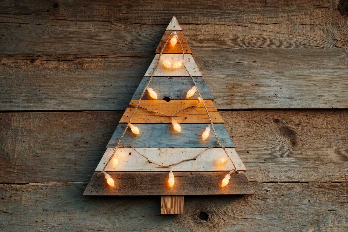 Albero di Natale stilizzato realizzato con assi di pallet dipinte in diverse tonalità naturali, decorato con luci calde e appeso su una parete di legno rustico.
