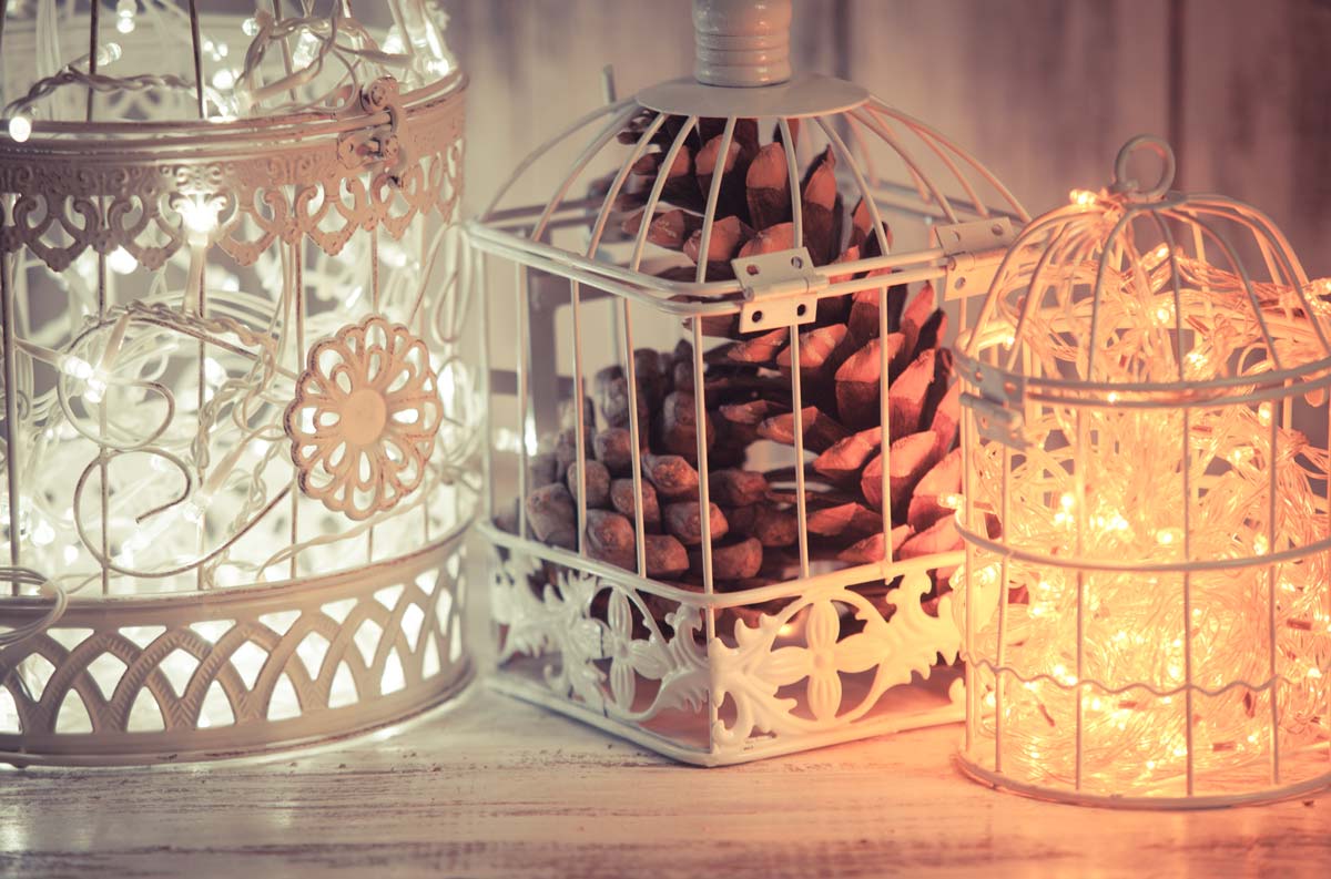 Tre lanterne shabby chic in metallo bianco, illuminate da lucine calde e decorate con dettagli floreali e una grande pigna, su sfondo in legno chiaro.