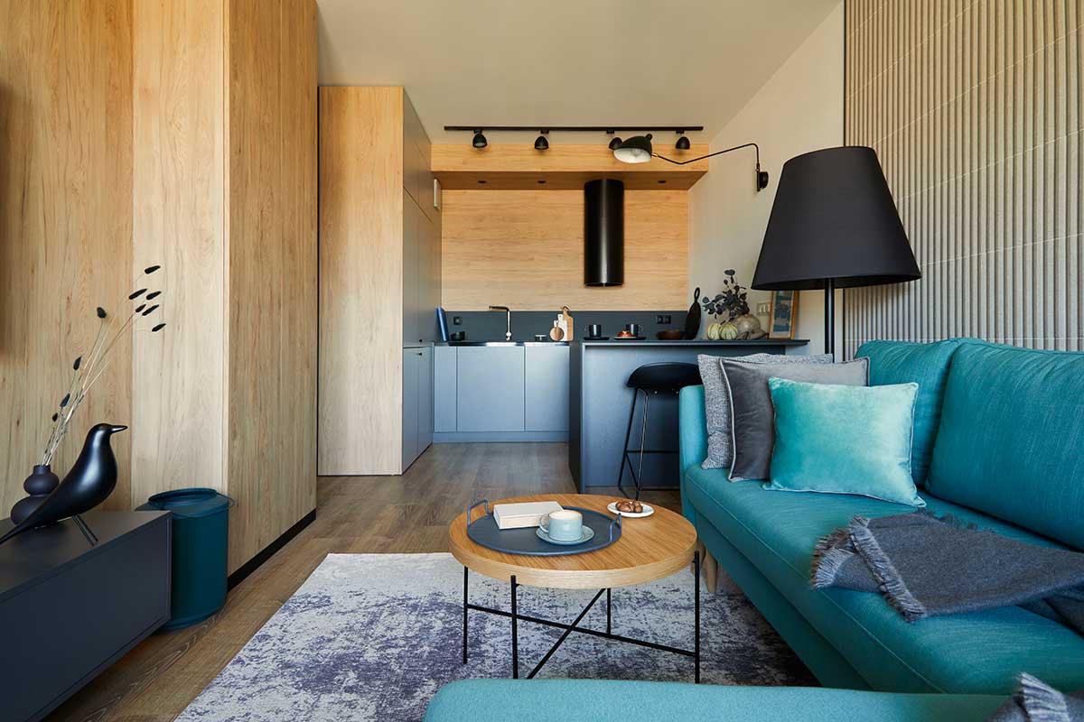 Piccolo open space moderno con cucina a vista, mobili in legno chiaro e divano turchese, tavolino rotondo, lampada nera e dettagli minimalisti. Ambiente luminoso e accogliente, perfetto esempio di soluzione intelligente per spazi ridotti.