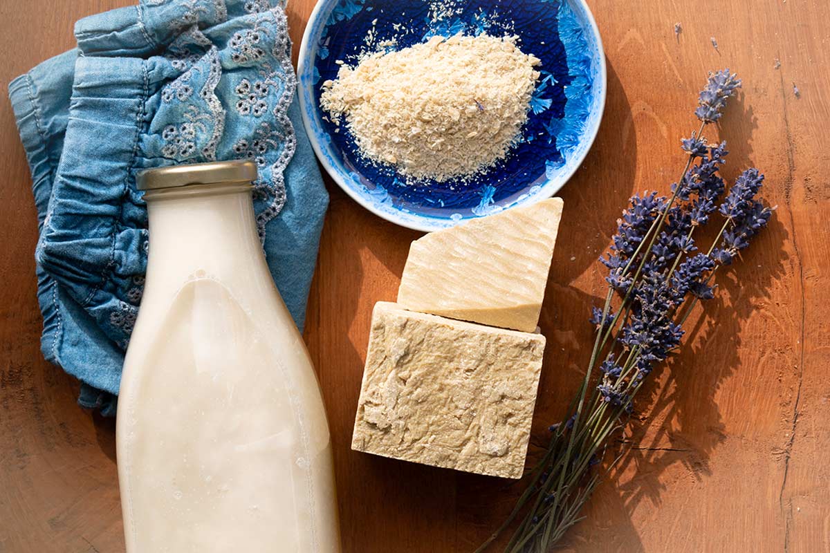 Blocchi di sapone di Marsiglia, scaglie in una ciotola blu, bottiglia di detersivo liquido artigianale, rametti di lavanda e tessuto ricamato blu su tavolo di legno, in un’atmosfera naturale e luminosa.