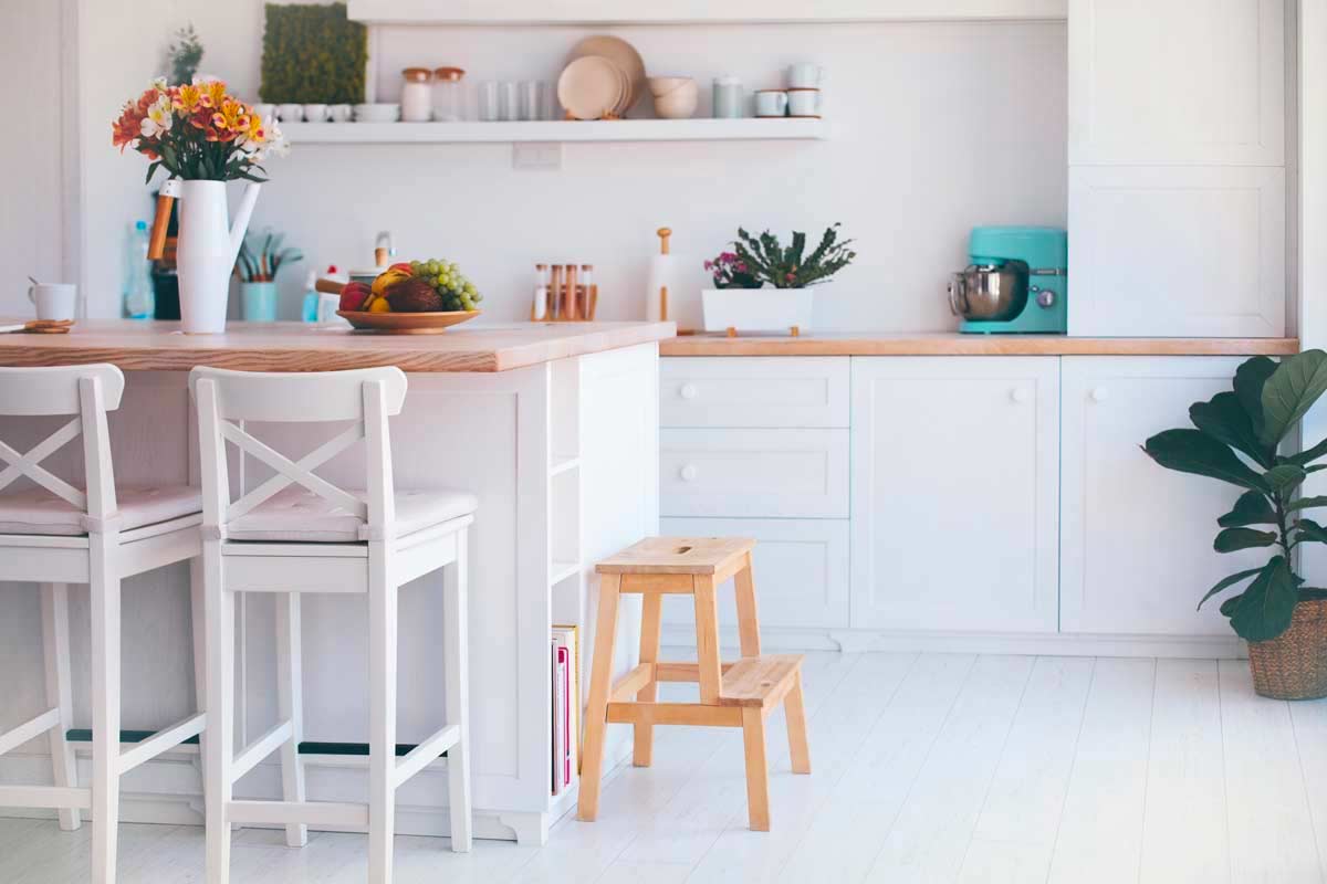 Cucina luminosa in stile nordico con mobili bianchi, piano in legno chiaro, sgabelli e sedie coordinate, dettagli naturali e accenti di colore verde e turchese. Sul tavolo centrale, un vaso di fiori freschi e una ciotola di frutta portano vitalità all’ambiente arioso e accogliente.