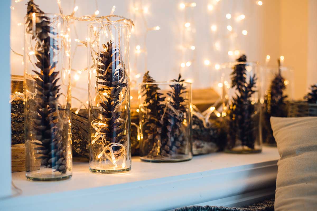 Vasi di vetro cilindrici riempiti con pigne allungate e fili di luci calde, disposti su una mensola insieme a tronchetti di legno, con sfondo di luci natalizie soffuse e atmosfera accogliente.