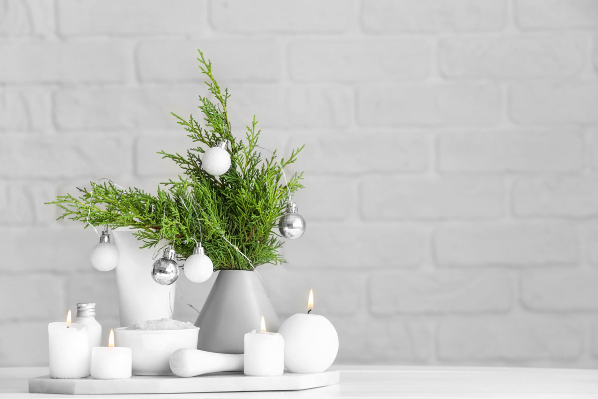Vaso grigio con rami verdi e palline bianche, candele accese su fondo chiaro. 
