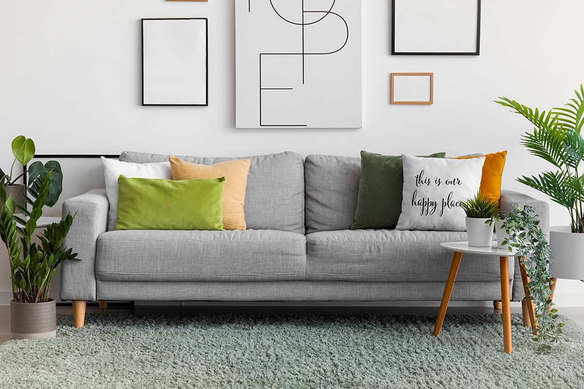 Divano grigio moderno con cuscini verdi, gialli e bianchi su tappeto verde chiaro, tavolino con piante e quadri minimalisti alle pareti; esempio di salotto luminoso con tappeto ben posizionato sotto le gambe anteriori del divano.