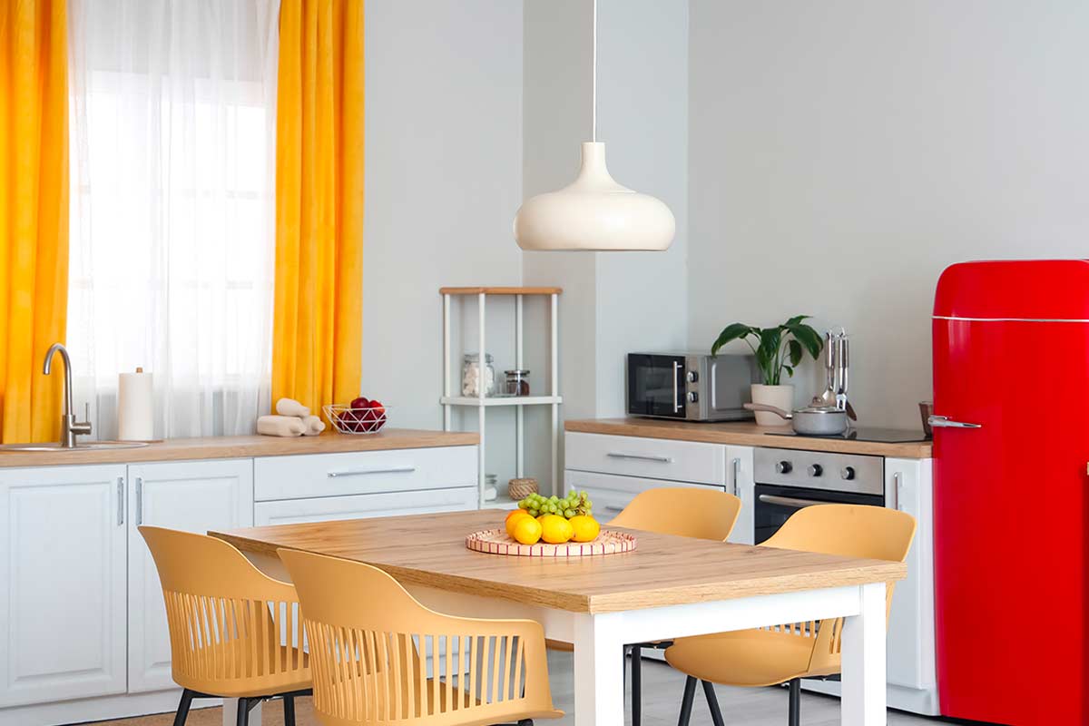Cucina luminosa in stile moderno con lampada a sospensione bianca posizionata sopra un tavolo quadrato in legno chiaro, circondato da quattro sedie gialle; sullo sfondo tende gialle e frigorifero rosso vintage.