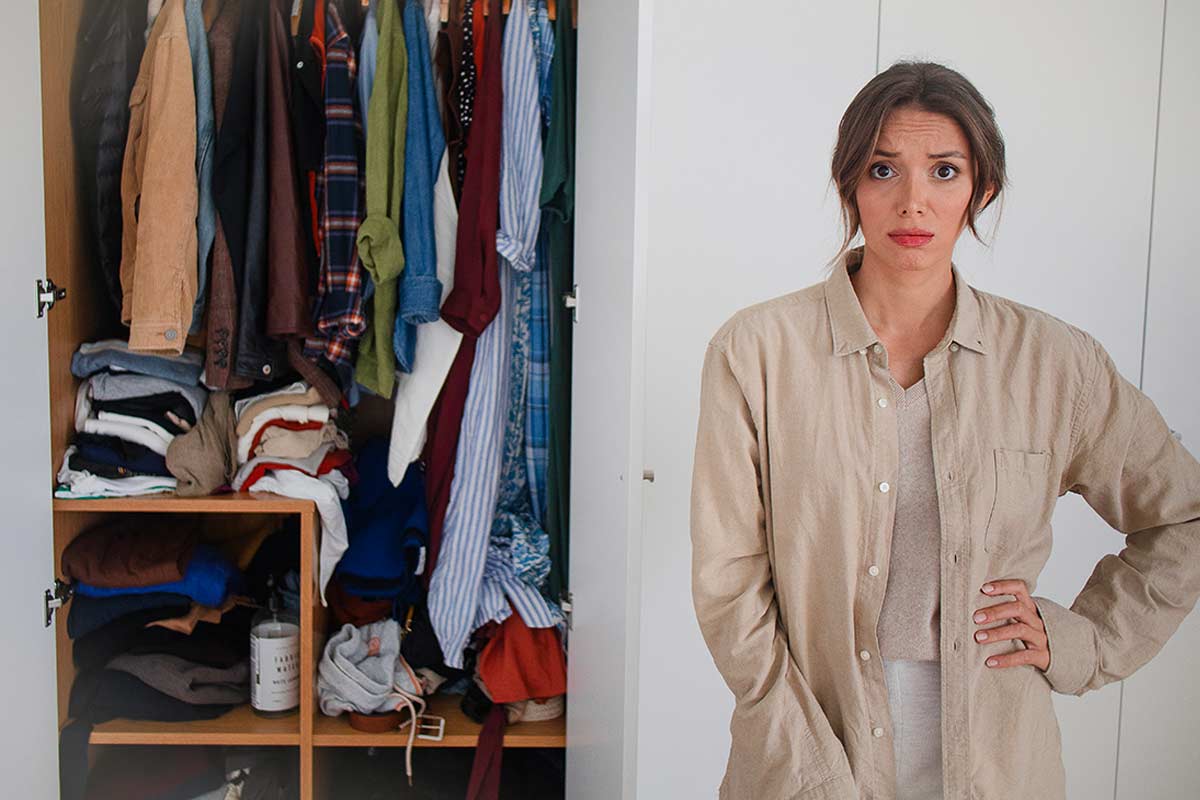 Donna davanti a un armadio disordinato, pieno di vestiti ammassati e ante spalancate, con espressione preoccupata, foto: © Lais - Stock adobe