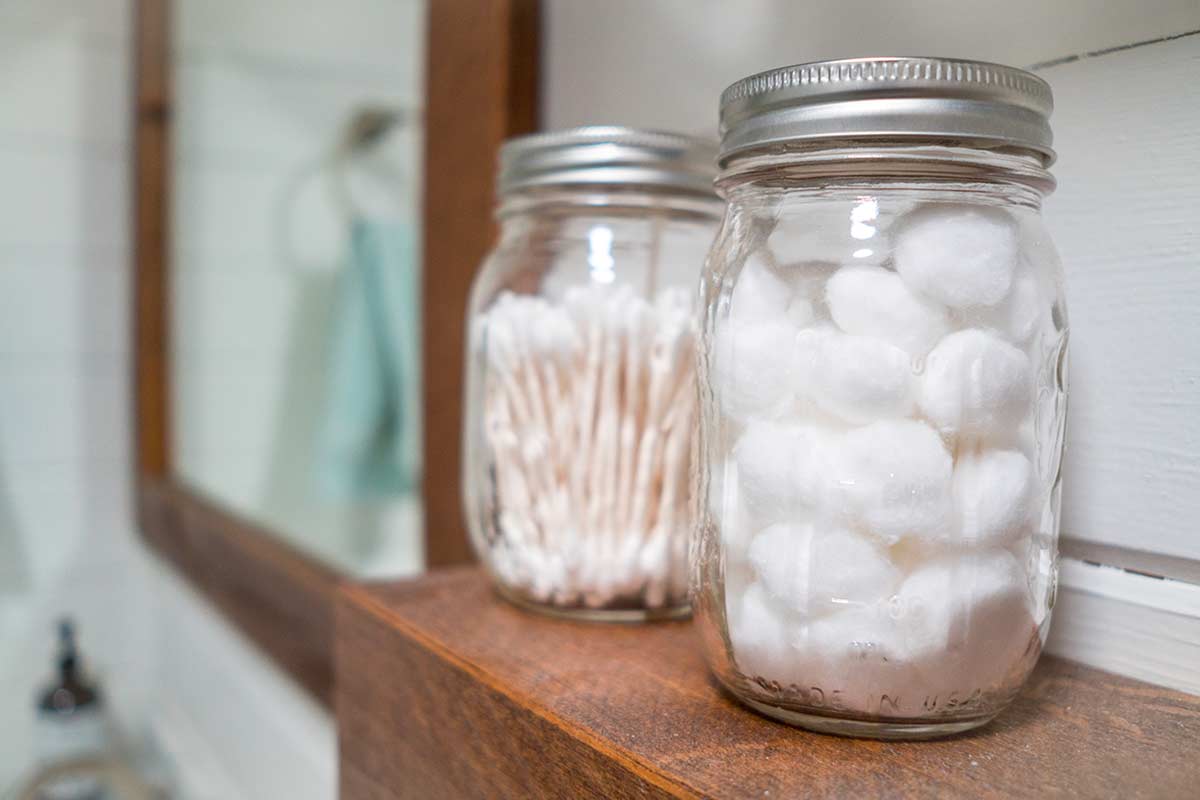 Barattoli di vetro trasparente con coperchio metallico, utilizzati per organizzare batuffoli di cotone e cotton fioc su una mensola in legno in bagno. La scena trasmette ordine, pulizia e un tocco di semplicità naturale.