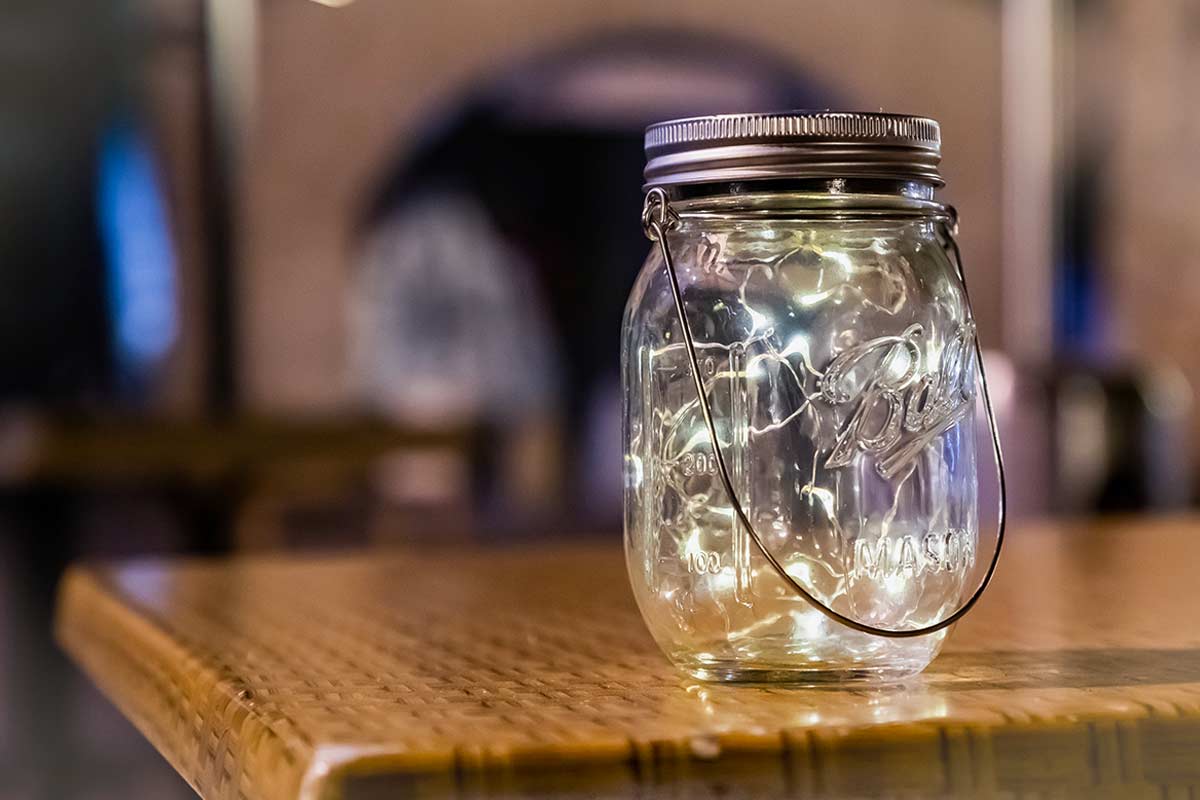 Barattolo di vetro con manico, usato come lanterna decorativa con luci LED all’interno, su tavolo in legno. Atmosfera calda e accogliente.