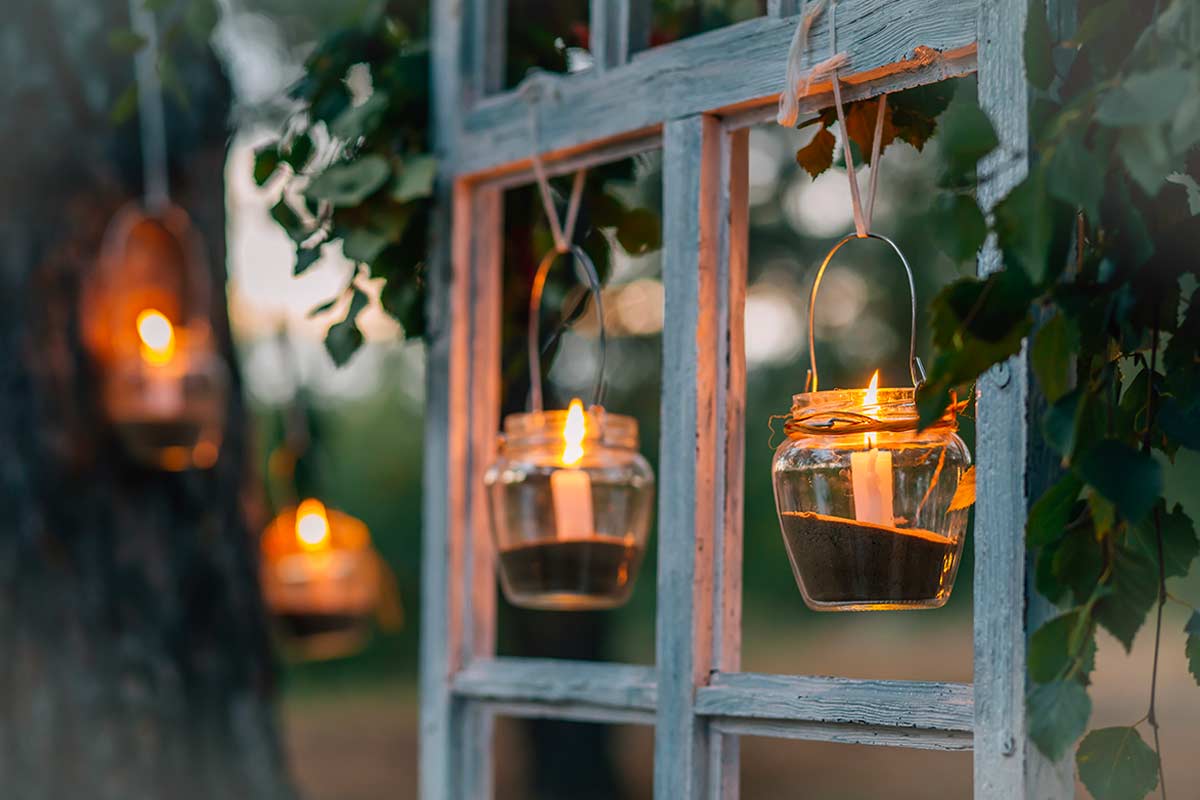 Barattoli di vetro appesi con candele accese, usati come lanterne su una finestra di legno all’aperto, tra foglie verdi. Atmosfera serale e suggestiva.