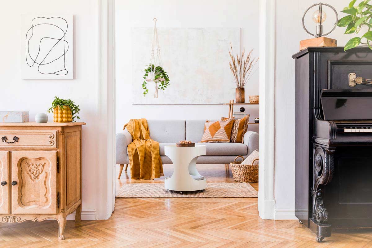 Soggiorno elegante in bianco e legno con parquet, divano grigio chiaro, dettagli ocra e beige, mobile vintage, piante e pianoforte nero in primo piano.