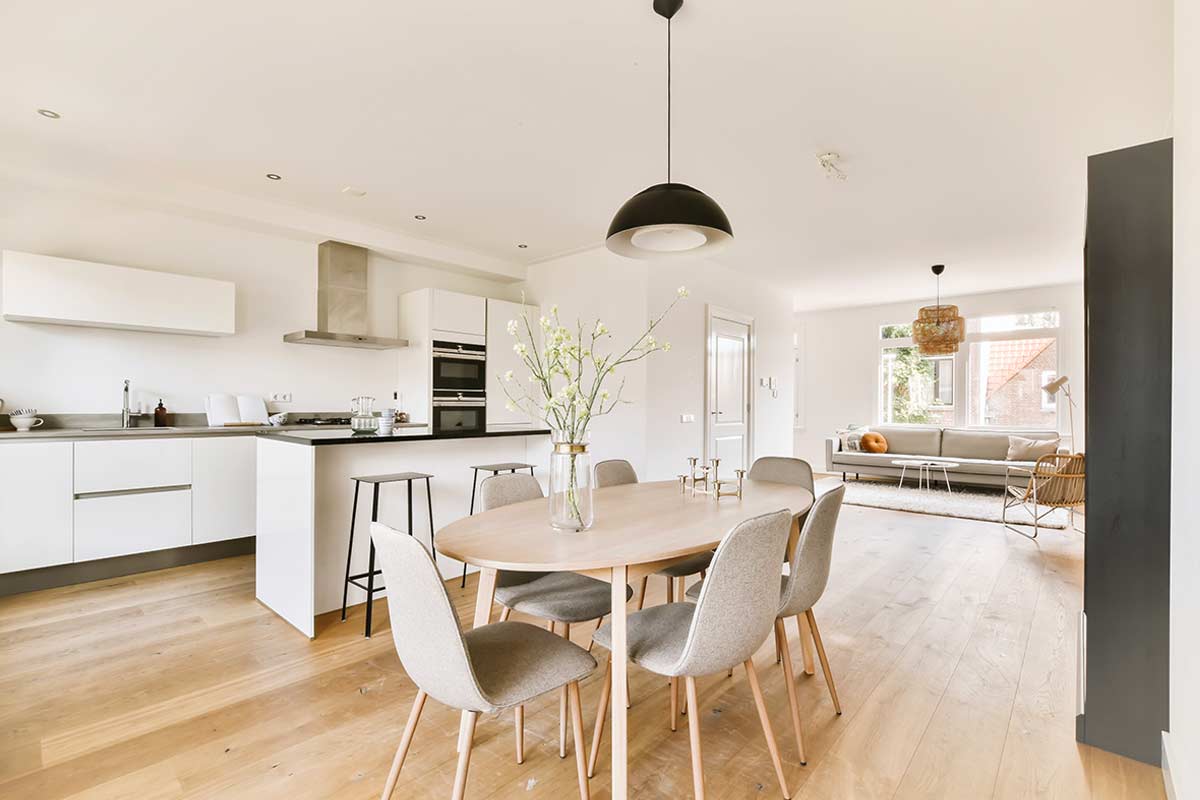 Cucina e zona pranzo open space in stile moderno con mobili bianchi, tavolo in legno chiaro, sedie beige, pavimento in parquet e soggiorno luminoso sullo sfondo.