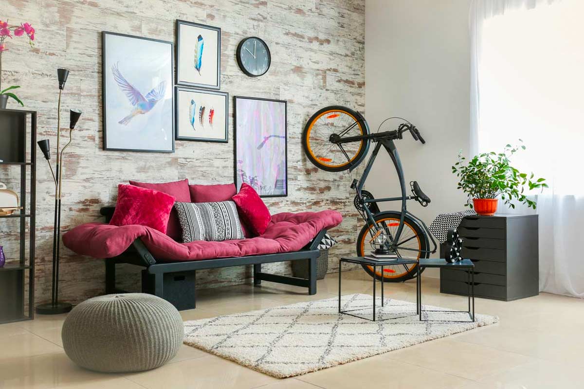 Divano futon bordeaux con cuscini chiari, parete in legno chiaro, bici appesa e dettagli colorati in un soggiorno moderno e luminoso.