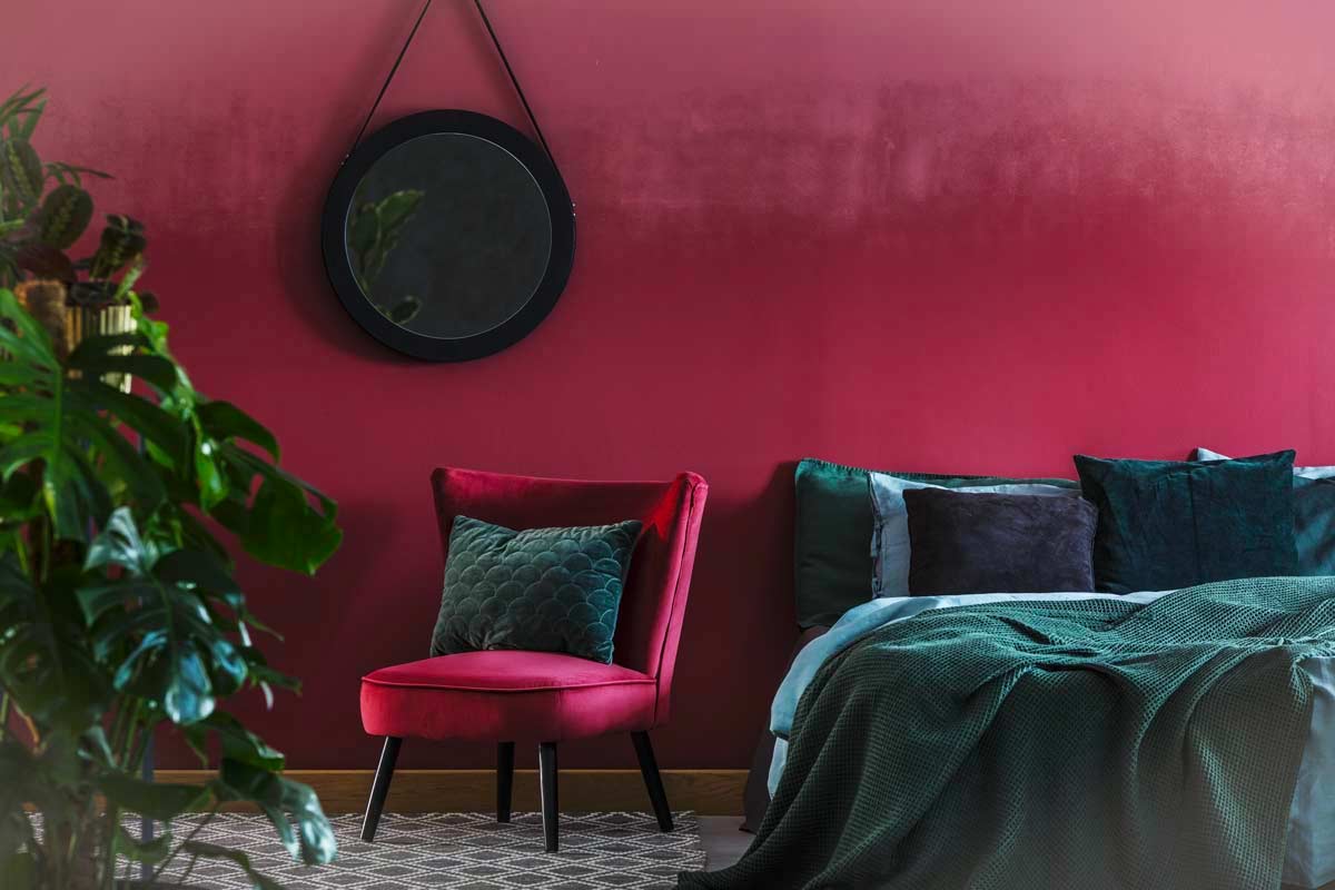Camera con parete bordeaux, poltroncina in velluto, cuscini e coperta verde scuro, pianta decorativa e specchio rotondo a parete.