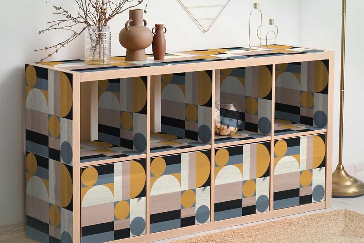 Mobile cubo modulare rivestito con carta da parati a motivi geometrici nei toni del giallo senape, blu scuro e beige. Sulla superficie, vasi decorativi in ceramica e vetro, in un ambiente minimal con dettagli in metallo dorato e tappeto in fibra naturale.