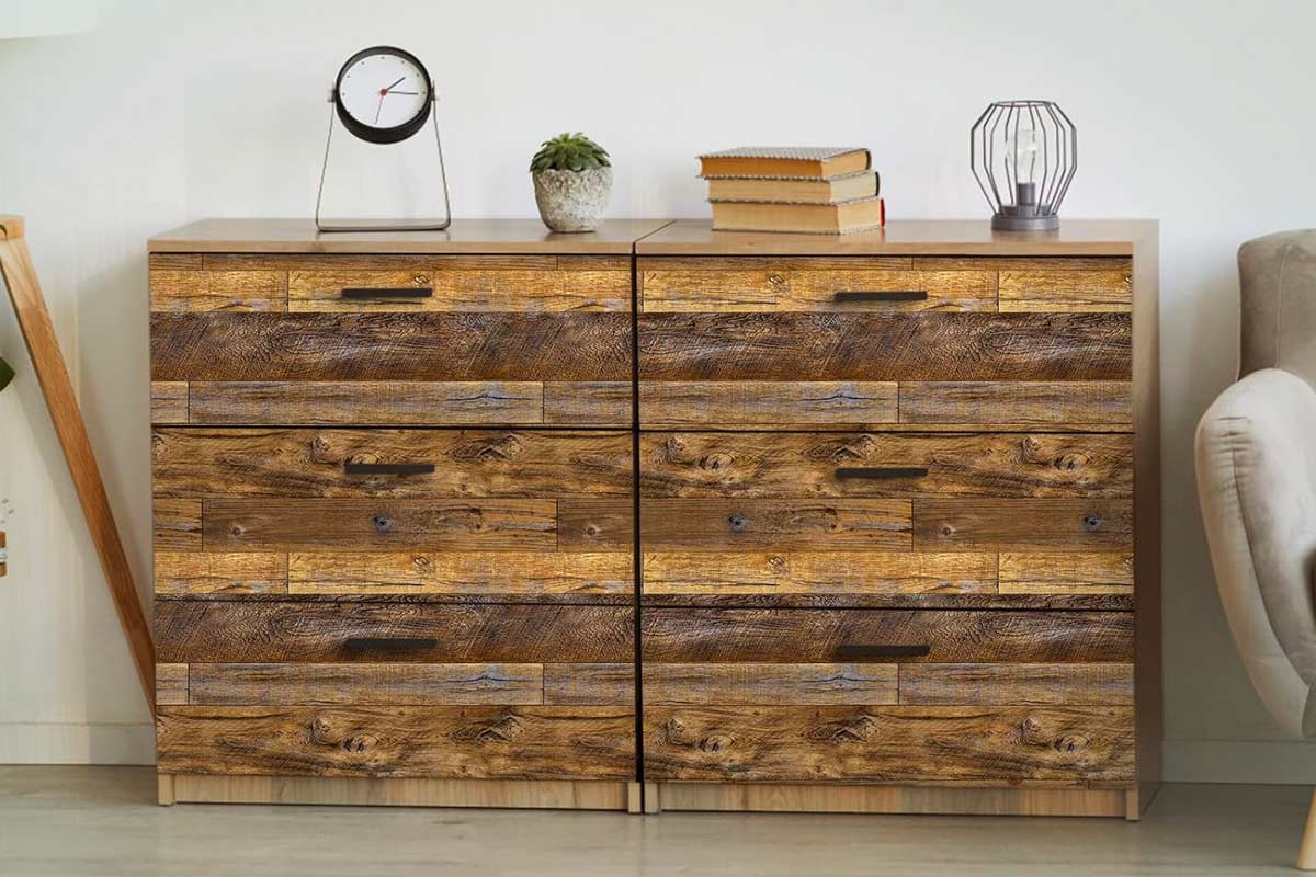 Cassettiera moderna rivestita con carta adesiva effetto legno vissuto, dalle venature marcate e toni caldi. Sul piano, orologio minimalista, pianta grassa, libri impilati e lampada decorativa in metallo, in un ambiente dal gusto naturale e accogliente.