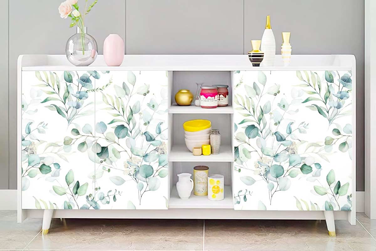 Mobile basso bianco rivestito con carta da parati a motivo botanico verde acqua, con ante decorate da foglie acquerellate. Sul piano vasi decorativi e al centro ripiani a giorno con ciotole e oggetti colorati, in un ambiente moderno e luminoso.