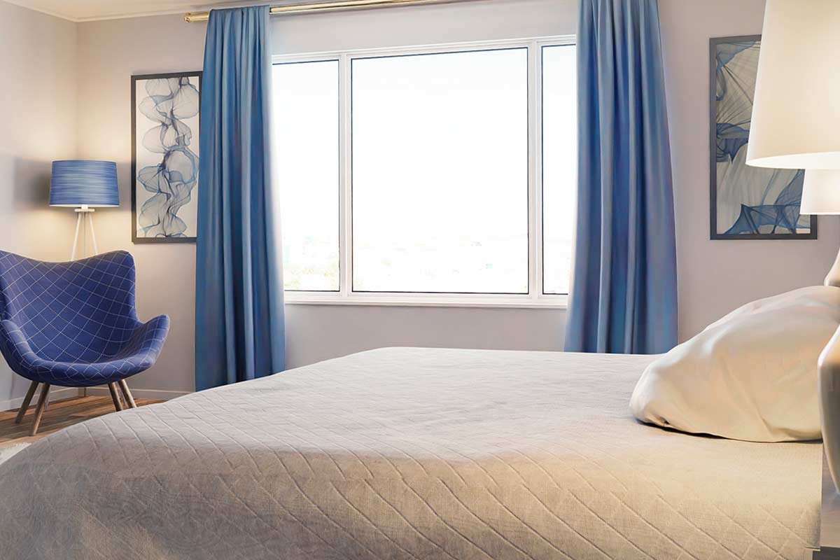 Camera da letto luminosa con ampie finestre, tende azzurre moderne, poltrona trapuntata blu e letto con copriletto grigio chiaro.