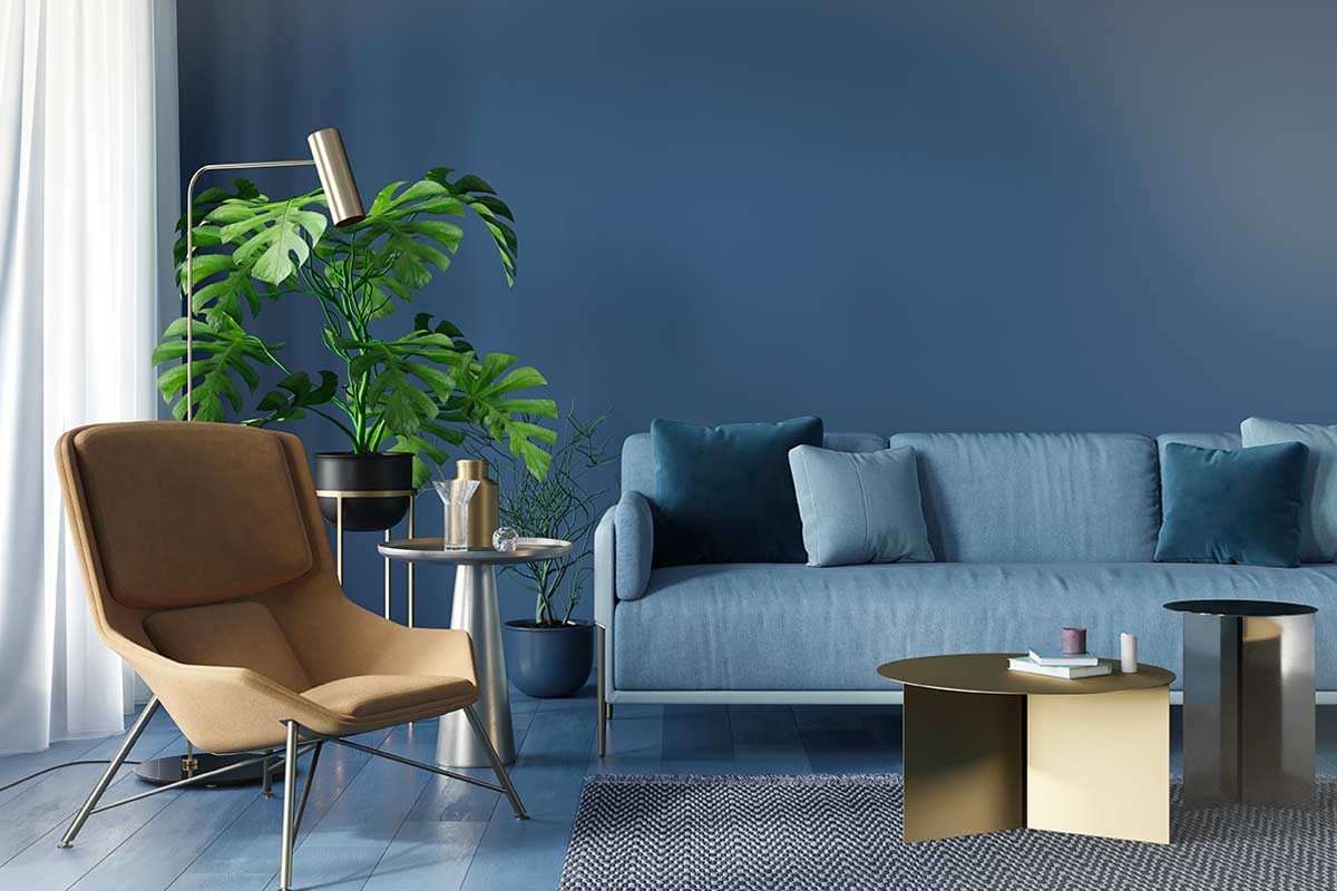 Salotto moderno con divano azzurro, parete blu, poltrona beige e piante verdi in vaso. Tavolini metallici e luce naturale filtrata da tende bianche.