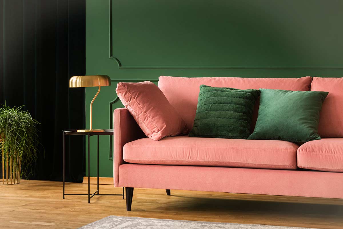 Divano rosa con cuscini verdi davanti a una parete verde scuro, lampada da tavolo dorata e pianta ornamentale su pavimento in legno chiaro.