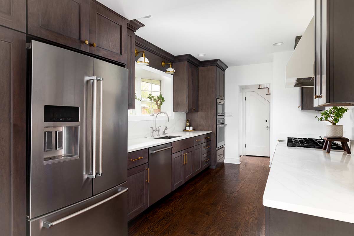 Cucina moderna con frigorifero in acciaio, mobili in legno scuro, pavimento in parquet caldo e dettagli eleganti. Luce naturale dalla finestra e piccoli oggetti verdi sul piano.