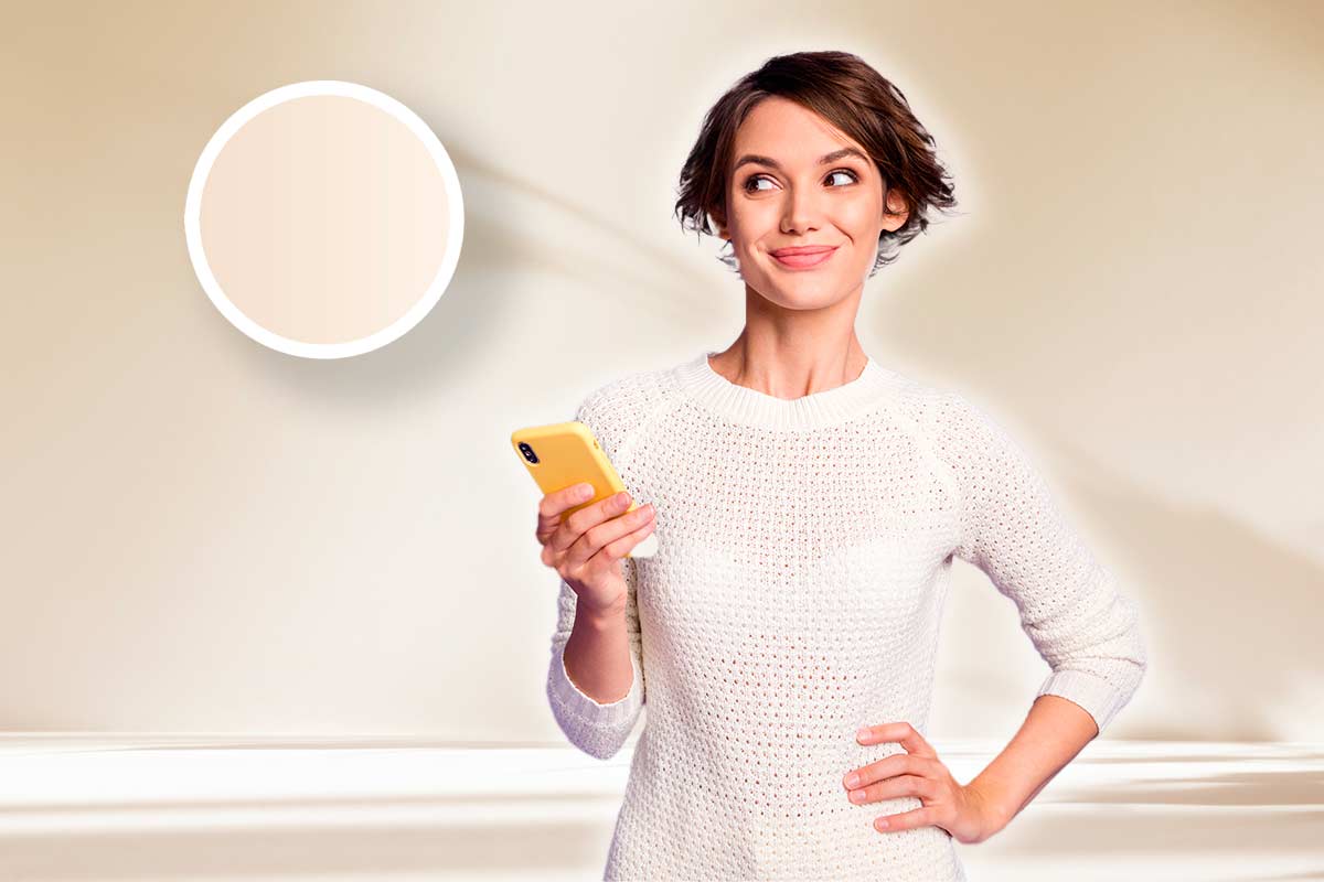 Donna sorridente con smartphone in mano su sfondo avorio luminoso, accanto a un campione circolare di colore avorio, simbolo della nuova tendenza del minimalismo caldo negli interni.