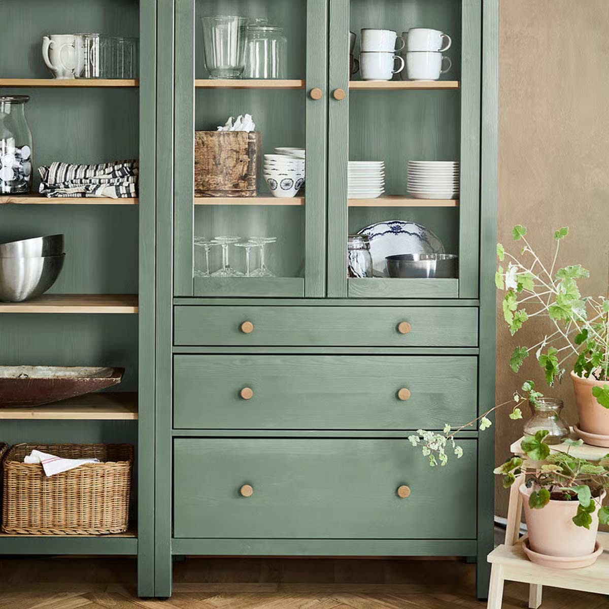 Credenza HEMNES IKEA verde salvia con cassetti, vetrina e ripiani, piatti e bicchieri esposti, piante in vaso a lato.