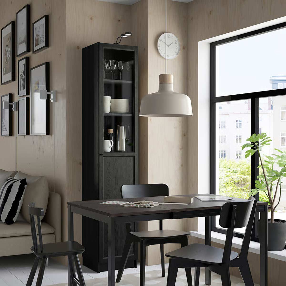 Vista di una zona pranzo moderna e luminosa con tavolo e sedie nere, vetrina BILLY/OXBERG nero effetto rovere di IKEA con ante a vetro, grandi finestre e dettagli di design minimalista.