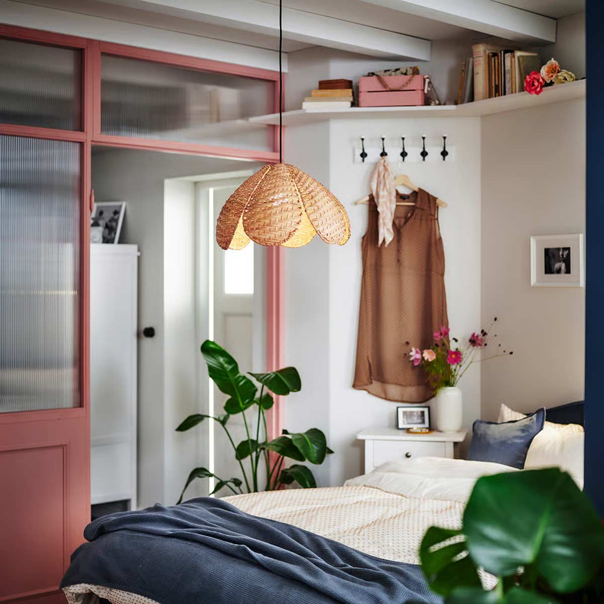 Vista d’insieme di una camera da letto moderna con la lampada SANDKORN di IKEA in rattan intrecciato sospesa sopra il letto, dettagli di arredi colorati e piante verdi.